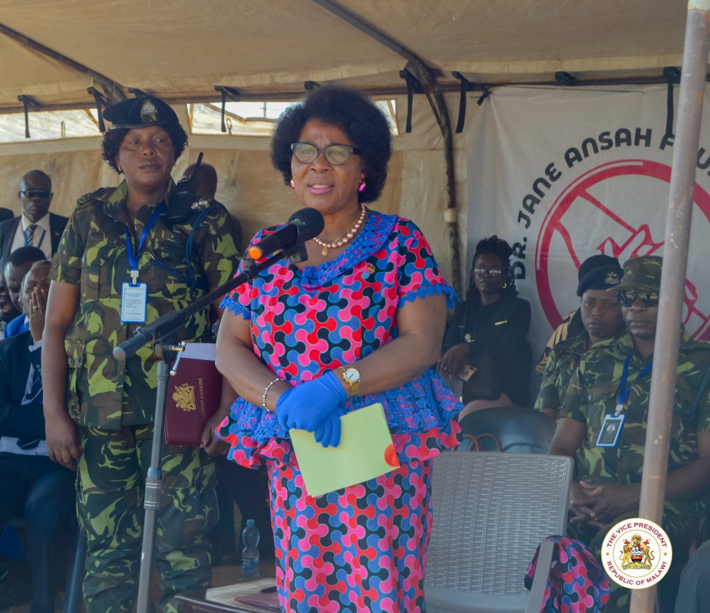 Dr. Jane Ansah, wearing blue gloves and a patterned dress, speaks from a podium with a microphone, flanked by an military aide. Behind her is a foundation banner.