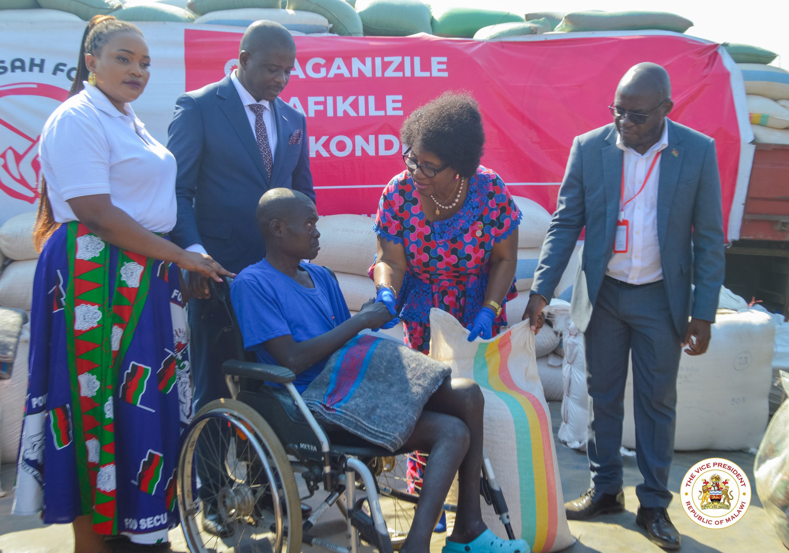 Dr. Jane Ansah, in a colorful patterned dress and blue gloves, is holding a large bag of relief aid, interacting with a young man in a wheelchair, surrounded by other foundation officials and a pile of supply sacks.